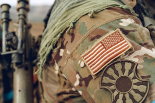 American flag patch on military camouflage uniform, representing patriotism and service, with tactical gear in the background.