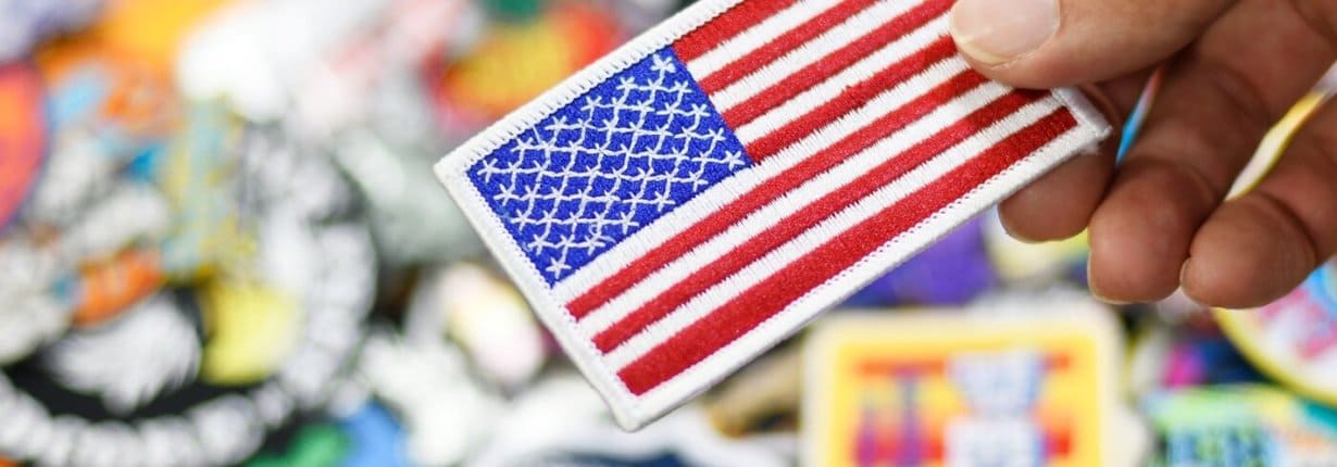 Hand holding an American flag embroidered patch with a colorful array of patches in the background, symbolizing patriotism and customization.