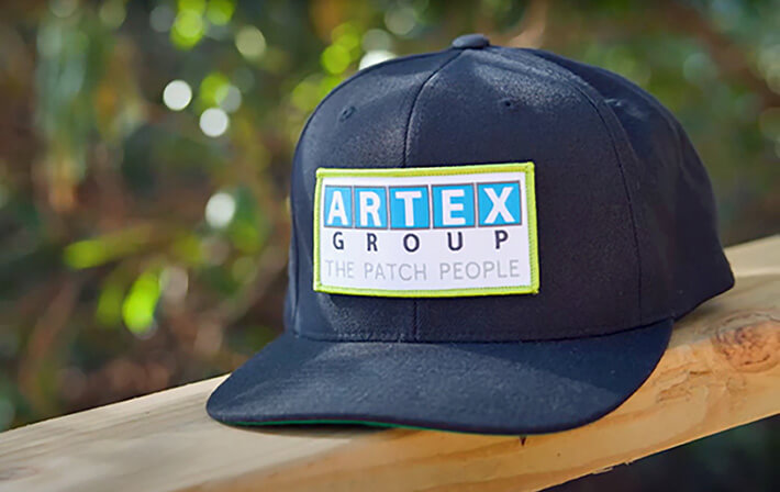 Navy baseball cap with lime-bordered Artex Group embroidered patch reading The Patch People on a wooden rail outdoors.